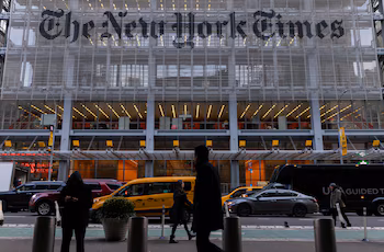 Вид спереди на здание New York Times с большой заметной вывеской. На улице внизу желтые такси, машины и пешеходы.