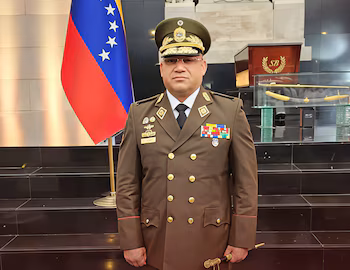 Генерал Карлос Александр Гомес Ларес, одетый в коричневую военную форму и награды, позирует перед флагом Венесуэлы.