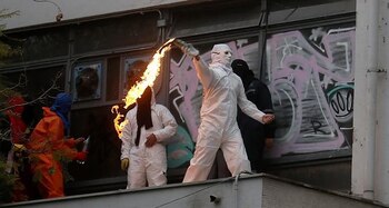 Несколько человек в масках, один в белом костюме и маске, выбрасывают пламя из предмета перед зданием с граффити на окнах.