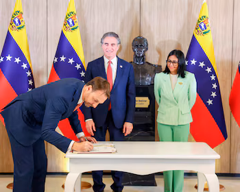 Мужчина в темно-синем костюме подписывает документ за белым столом в гостиной в окружении двух человек и венесуэльских флагов.