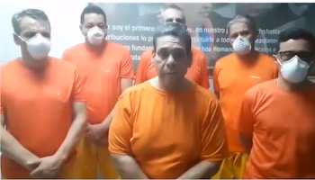 Шесть мужчин, пятеро в белых масках и центральный без масок, носят оранжевые футболки на фоне с частично видимым текстом.