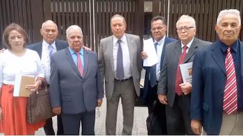 Группа из семи адвокатов, шести мужчин и одной женщины, стоит перед зданием. Большинство из них одеты в формальную одежду, а некоторые держат в руках документы.