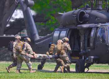 Пять солдат в камуфляжной форме несут человека на носилках к серому военному вертолету с открытой дверью на травяном поле
