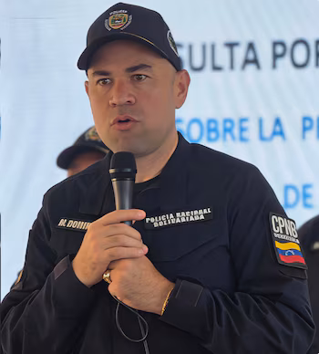 Крупный план Мигеля Домингеса, заместителя директора PNB, в темной форме, с микрофоном в руках. Фоном является экран с размытым текстом.