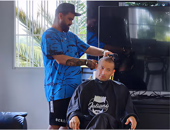 Парикмахер в кепке и синей рубашке стрижет волосы Черемо, который носит черную накидку с логотипом Джулиано, в интерьере с окном и телевизором.