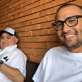 Сидят двое улыбающихся мужчин; старший в кепке и белой рубашке, а молодой в очках и белой футболке. кирпичная стена фон