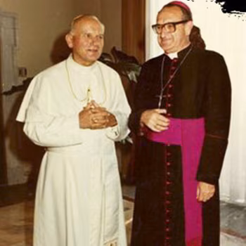 Епископ Хуан Хосе Херарди Конедера в темной рясе и пурпурном поясе стоит рядом с Папой Иоанном Павлом II, одетым в белое, оба улыбаются.