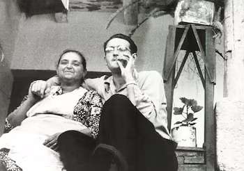 Черно-белое фото молодого человека в очках и пожилой женщины, сидящей и улыбающейся на фоне горшечных растений.