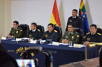 Пять офицеров в форме сидят за синим столом с микрофонами; офицер, стоящий позади. Видны боливийские флаги и детали «Боливийской Армады».