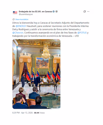 Мужчина в темном костюме подписывает документы за столом с флагами Венесуэлы. Другие посетители наблюдают за происходящим в формальной комнате с люстрой и картиной всадника.