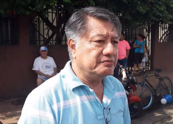 Портрет леди М, женщины с короткими каштановыми волосами и в очках, в полосатой футболке и золотом кресте, сидящей на открытом воздухе