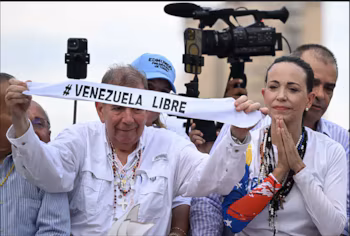Мужчина и женщина держат белый баннер с надписью «#VENEZUELA LIBRE». На заднем плане оператор с телекамерой и мобильным телефоном.