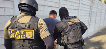 Трое полицейских сопровождают мужчину в наручниках, красной рубашке и серых шортах мимо полицейской машины и деревьев.