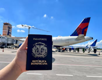 Рука держит черный доминиканский паспорт с золотым щитом перед красно-синим хвостом самолета Delta на взлетной полосе в международном аэропорту Сибао.