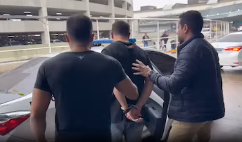 Двое мужчин сопровождают мужчину в наручниках к белой машине с открытой дверью на многоуровневой парковке на заднем плане.