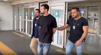 Мужчину в черной рубашке и джинсах перед стеклянными дверями сопровождают двое полицейских в темных футболках и значках.