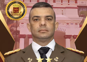 Бюст портрет мужчины в коричневой военной форме, белой рубашке, черном галстуке. Золотые значки на лацканах и плечах. Золотая эмблема. Архитектурный фон