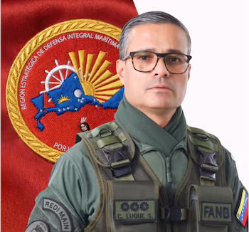 Портрет мужчины в очках, зеленой военной форме, тактическом жилете, заплатах "РЕДИ ГЛАВНАЯ", "ФАНБ", "К. ЛУКЕ С." и флаг Венесуэлы. Красный фон с военно-морской эмблемой