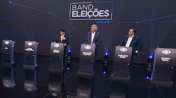 Трое мужчин стоят за пюпитрами на черном фоне с надписью Band Eleições 2026 в освещенной студии.