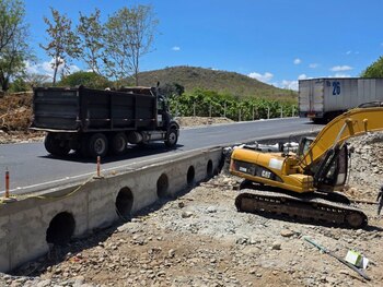 Желтый экскаватор Caterpillar рядом с бетонной стеной с круглыми водопропускными трубами и черным грузовиком, едущим по дороге