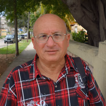 Крупный план лысого мужчины средних лет в очках и красно-черной клетчатой ​​рубашке, на фоне тропинки и зеленых деревьев.