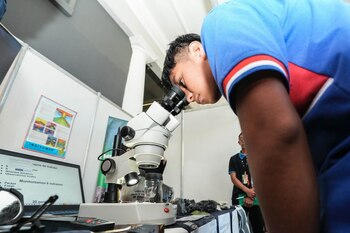 Наклонившийся молодой человек смотрит в оптический микроскоп перед столом с камнями, колбами и ноутбуком.