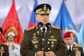 Владимир Падрино Лопес носит военную форму с фуражкой и знаками различия и говорит в два микрофона с флагами позади.